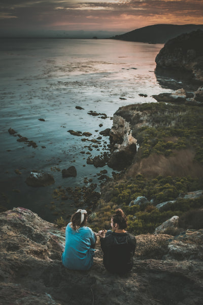Two girls sit together on the top of a cliff overlooking the seaside at sunset