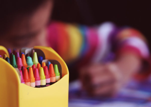 The Ultimate Back to School List for 2021. Close up of a box of crayons with a girl drawing with one in the background.