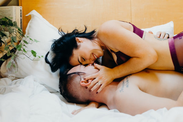 Why the Link Between Sex and Emotional Connection Is So Important: Couple smiling at each other as the girl ups the guy's face in her hands as they lie in bed.