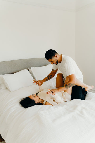 Why You Should Have a Sexual Bucket List: Couple sharing laughs together as they lie in bed with white sheets.