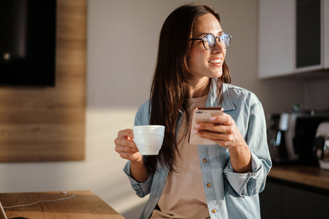 Happy charming woman using smartphone and drinking coffee at home kitchen