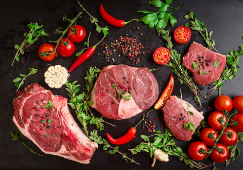 Different types of steaks set. Assorted raw meat on a black chalk board background. Rib eye steak on the bone, veal shank (ossobuco), fillet with cherry tomatoes, hot pepper and herbs.