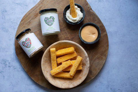 Large wooden board in the centre of the photo; this is topped with a smaller wooden bowl of crisp thick cut golden polenta chips. Above the wooden bowl is a smaller bowl of red chilli mayonnaise, with 2 jars of Lucy's vegan mayonnaise beside.