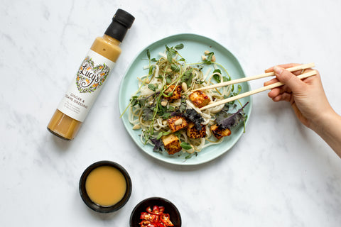 Plate of noodles with tofu on top greens, chilli and chopsticks tucking in. On the left, there is a bottle of Lucy's ginger and sesame, with 2 smaller bowls next to it of dressing and chopped up chilli. 