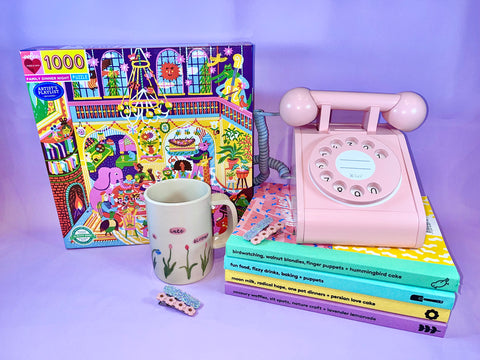 Photo of a puzzle box, a mug, 4 hair clips, a pink toy phone and a stack of magazines on a lilac background