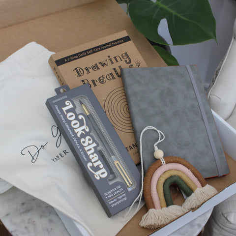 Open subscription box filled with tote bag, Drawing Breaths book, grey journal, graphite pencil and macrame rainbow