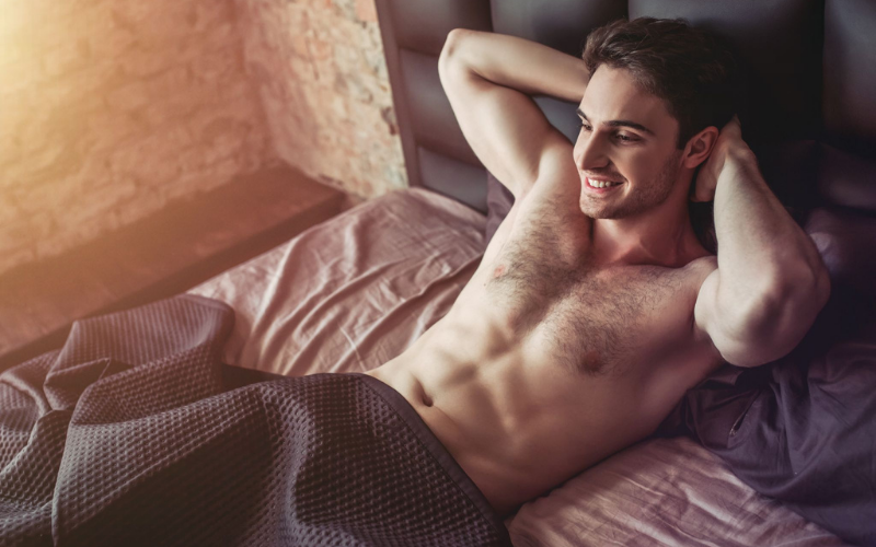 Man laying in bed shirtless with hands behind his head and a big smile