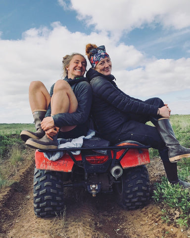 Elma and her friend wearing a headband and Xtratufs while sitting back to back on the back of a four-wheeler.