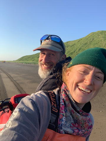 Elma wearing coveralls, a green knit hat, and Skida brand bandana while riding on a four wheeler with her friend in Bristol Bay.