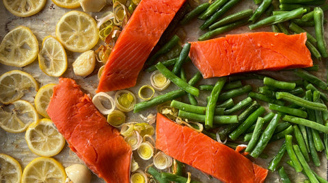 Bright red sockeye salmon portions nestled among a baking sheet of yellow lemon slices and green vegetables.