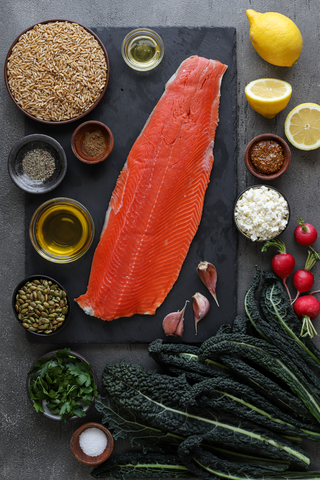 Salmon fillet on a black cutting board surrounded by lemon, kamut, feta, kale, radishes, herbs and spices