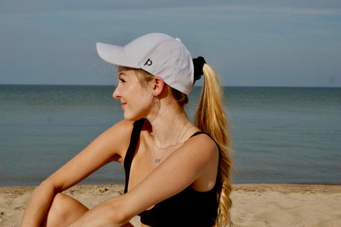 Girl sitting on beach with white ponyback ponytail hat on smiling to the side with water and sand behind her