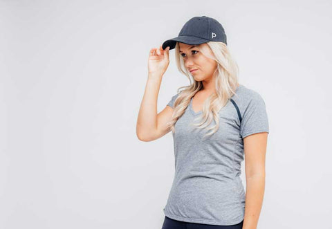 Woman wearing black ponyback hat with hair down and grey shirt tipping hat down and looking away with grey background indoors