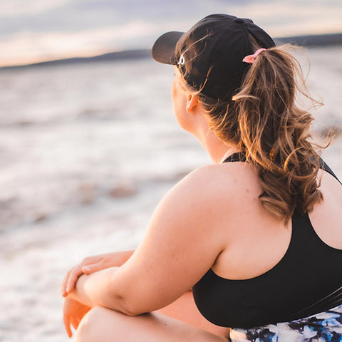 Woman sitting with waves in the background wearing a black Ponyback ponytail hat