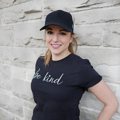 Girl with black ponytail hat on and black t-shirt that says be kind smiling at camera with grey brick wall behind her