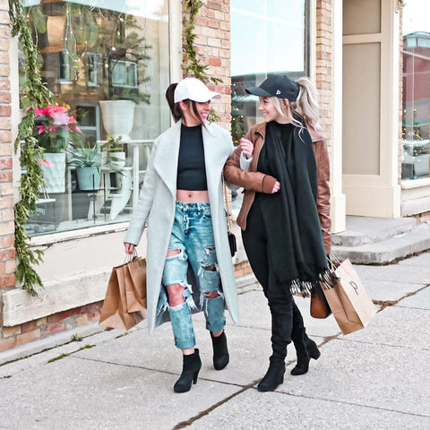 Two girls walking arm in arm down a street with stores holding shopping bags and wearing ponytail hats