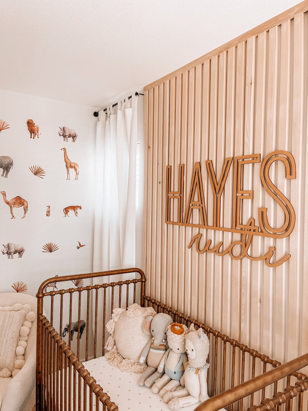 Nursery decorated with safari animal wall decals, featuring a wooden crib, plush toys, and a wooden slat accent wall with the name 'Hayes' in large wooden letters.