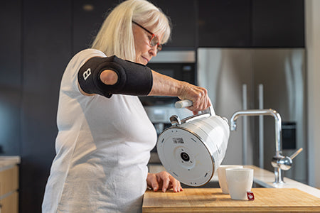 Elderly Woman pouring a kettle while wearing a Bio Magnetic Elbow Support in Black.