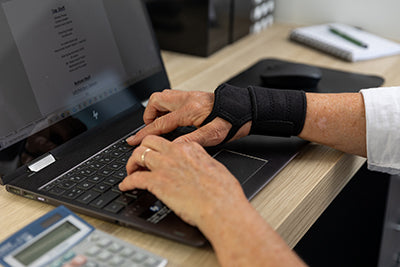 Woman's hands typing at a laptop while wearing a BioMagnetic Wrist Support in black on her right hand. 