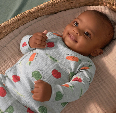 Baby boy laying down in woven bassinet wearing a gray outfit with vegetables