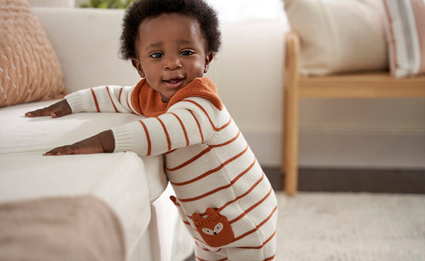 What a cutie! This baby boy is ready for anything in his trendy brown and white outfit, standing confidently by the couch.