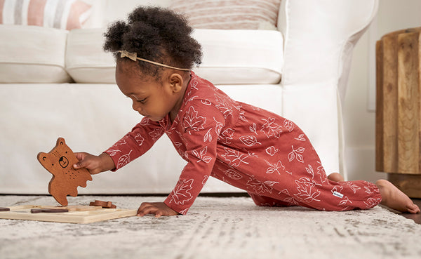 A cute baby girl in an orange outfit happily crawls on the floor, engrossed in playing with a large wooden puzzle piece.