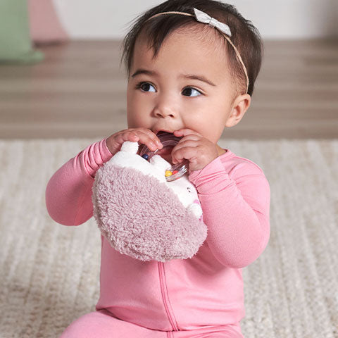 Baby girl in pink outfit teething on hedgehog soft toy