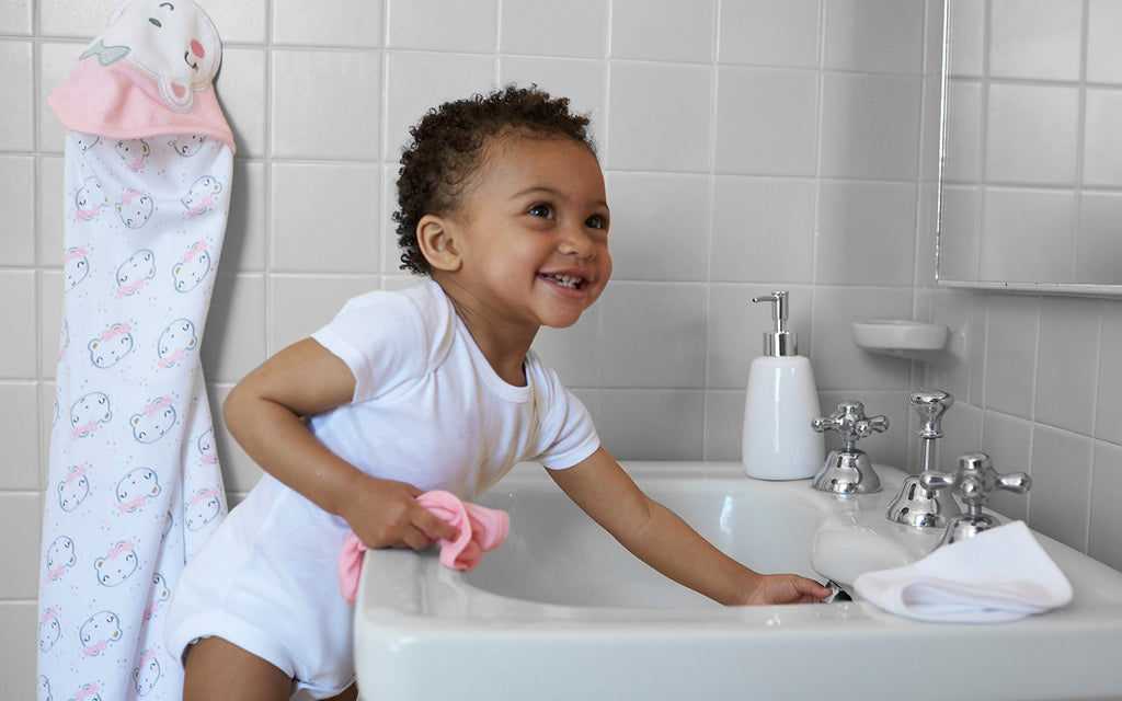 toddler in white bodysuit leans over sink holding washcloths