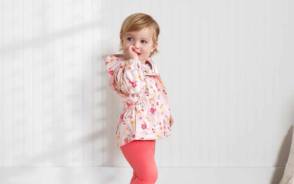 baby girl stands in against white wall wearing pink legging and matching jacket