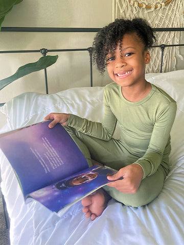 Maliyah, Mina's daughter, sitting down and smiling at the camera while holding the "Dancing on the Moon" book.