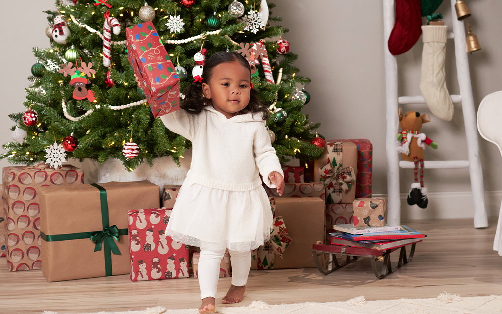 little girl in white dress holding red present and walking away from christmas tree