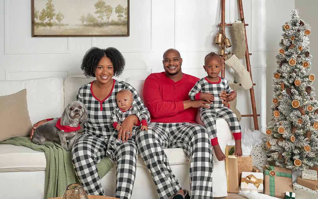 a family, mom and dad with baby and toddler, sit on couch with their dog next christmas tree. they are wearing black and white plaid pajamas with red accents. dog wears matching red bandana.