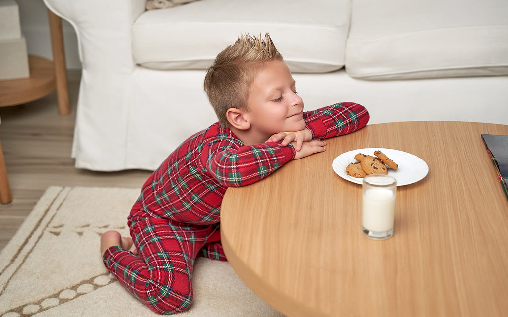 a little boy wearing red plaid pajamas sits in front of plate of cookies left for santa with a mischievous look on his face