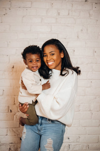 mother holds son while standing in front of white brick wall, both smiling