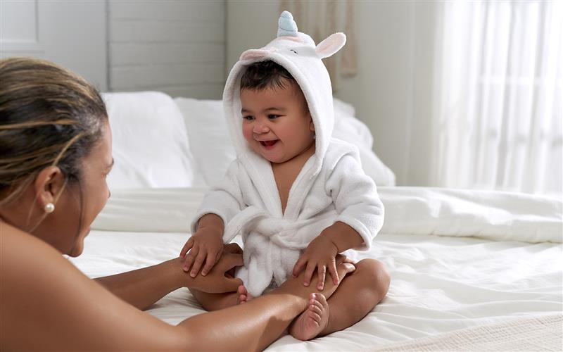 baby sitting on bed in white unicorn hooded towel looking at mother