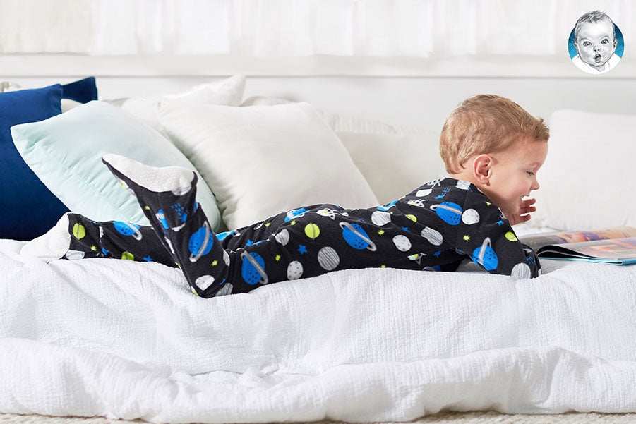 A toddler wearing footed pajamas is peacefully engrossed in reading a book while lying on a bed.