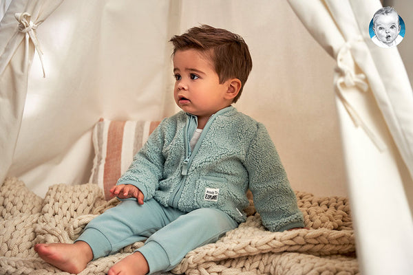 Small boy in light blue nubby zipper jacket sitting on a light colored couch.