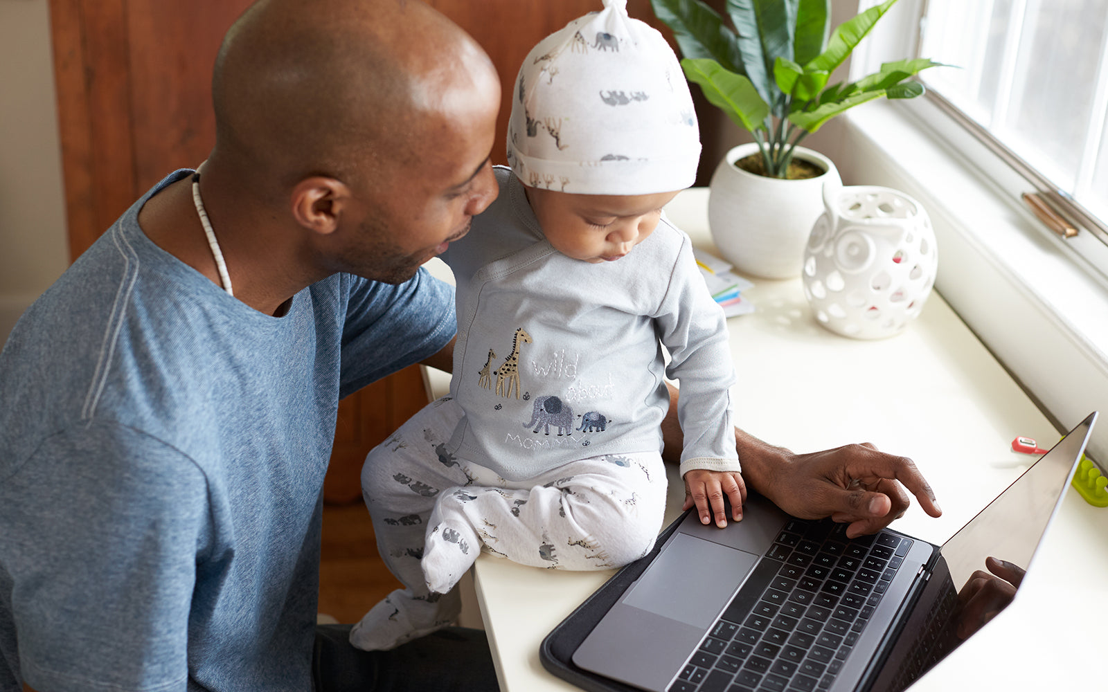 baby sits on desk next to laptop with dad watching carefully