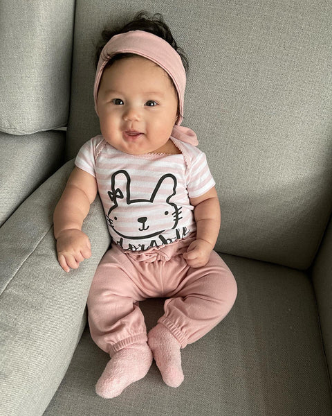 Baby girl wearing bunny Onesies bodysuit, pink pants and socks sits in chair smiling