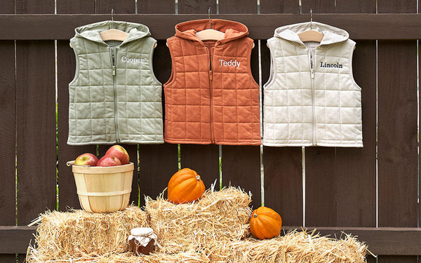 Fall toddler vest jackets hanging up on a brown picket fence in front of fall decor.