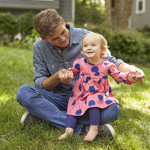 baby girl in pink and blue dress sits on father's lap outside