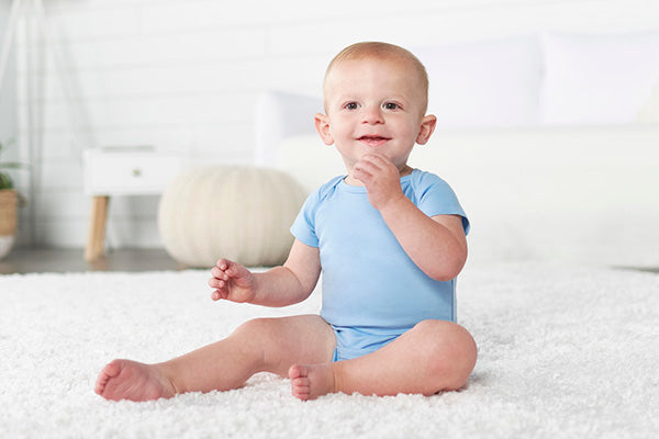 A sweet little baby sitting on a white rug, enjoying a moment with his hands in his mouth.