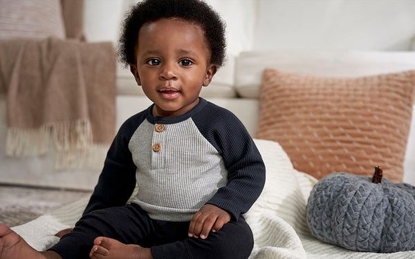 Young baby boy in two toned blue and grey shirt sitting next to a grey crochet pumpkin.