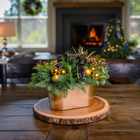 Centerpiece made of noble fir, cedar, and pine with Australian pine cones, faux gold berry clusters, gold ball clusters, a spray of gold stars, a black velvet bow with gold flecks and edging, and an oval-shaped metallic gold plastic container on a wooden table