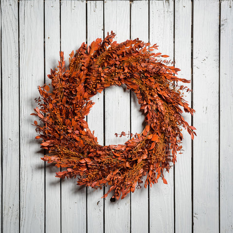 22" wreath made of orange myrtle, eucalyptus, and flax pods on a white wood fence background.