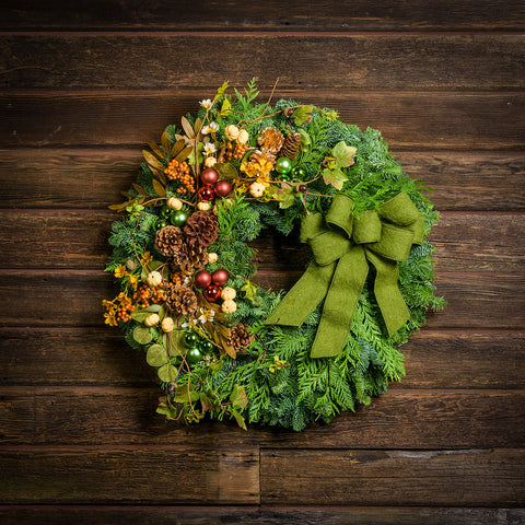 A holiday wreath of noble fir and western red cedar with faux pumpkins, faux acorns, sage accents, leaf and berry accents, 2 copper ball clusters, 2 bright green ball clusters, 3 gold pinecones, 4 Australian pinecones, 1 ponderosa pinecone, and a brushed green linen bow on a wood background.