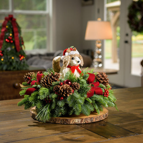 A holiday centerpiece of noble fir, white pine, incense cedar, 3 small pinecone and red berry decorations, 3 ponderosa pinecones, 3 red berry clusters, 3 red velvet bows, and a sisal Christmas dog decoration sitting on a wood round on a table.