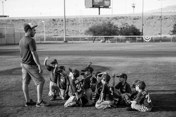 Black and white photo of a coach standing and talking to his team of little kids sitting in the grass