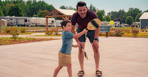 A man and his son at a park while the dad helps his son learn pickle ball