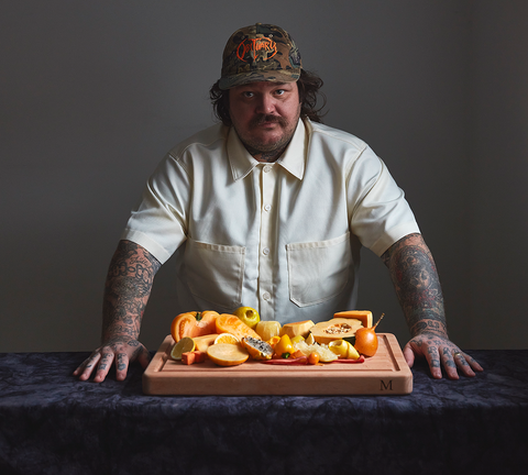 Matty Matheson in an off white button up and camouflage baseball hat with orange Obituary stitching, standing behind countertop with cut up orange food, atop the new Matheson Cookware Cutting Board. 